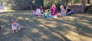 Na pierwszym planie widać dziewczynkę raczkującą w ogrodzie. w tle widać rozmawiających rodziców i opiekunki.