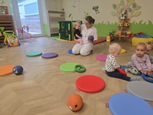 Ciocia bawi się piłką z dziećmi. Wokół dzieci na podłodze są kolorowe krężki.
