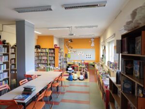 Widok środka biblioteki i pani czytającej dzieciom książkę na dywanie