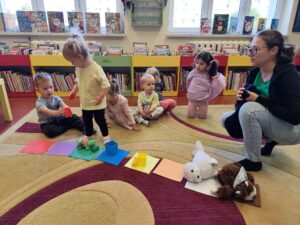 Dzieci siedzą na dywanie w bibliotece i dopasowują przedmioty o odpowiednim kolorze do kartek które są rozłożone przed nimi na dywanie