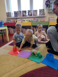 Dzieci siedzą na dywanie w bibliotece, chłopiec kładzie czerwony liść na czerwonej kartce