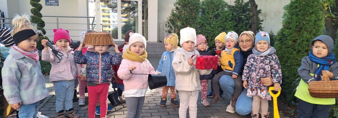 Dzieci stoją przed żłobkiem z koszyczkami oraz narzędziami ogrodniczymi