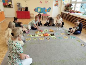 Dzieci wraz z rodzicami siedzą na dywanie, opiekunka pokazuje dary jesieni
