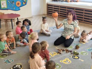 Dzieci siedzą na dywanie a opiekunka pokazuje im dynię