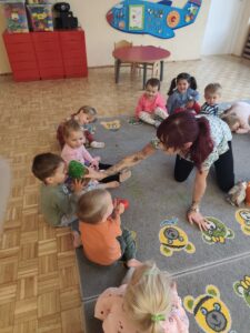 Dzieci siedzą na dywanie a opiekunka pokazuje im pluszowe drzewo