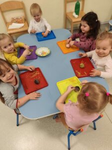 Dziewczynki przy stole wtykają makaron spaghetti w łupinkę orzecha oklejoną plasteliną