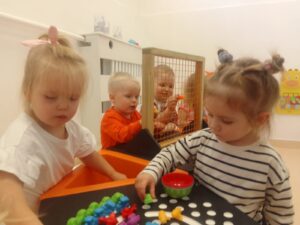 Dzieci układają tęczę z kolorowych ksztatków na stole sensorycznym, w tle dzieci wkładają kształtki przez siatkę
