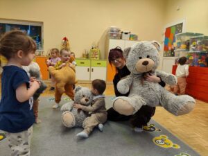 Opiekunka trzyma dużego Misia, w tle dzieci ze swoimi maskotkami