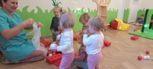 Ciocia bawi się z dziećmi chustkami. W tle widać dzieci bawiące się piłeczkami.