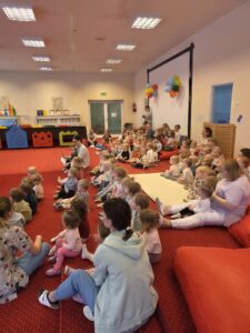 Dzieci siedzące na czerwonym dywanie w sali żłobka oglądają przedstawienie teatralne. Przed nimi ustawiona jest niewielka scena z dekoracją przedstawiającą statek na morzu, zasłonięta po bokach granatowymi kotarami. W tle widać kolorowe meble i zabawki, a opiekunki siedzą razem z dziećmi, wspólnie uczestnicząc w wydarzeniu. Atmosfera skupienia i zaciekawienia.