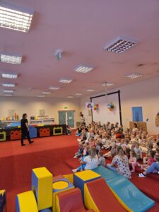 Grupa małych dzieci siedzi blisko siebie na czerwonym dywanie w sali żłobka i z dużym skupieniem ogląda przedstawienie poza kadrem. W tle widać opiekunki siedzące razem z dziećmi. Na twarzach maluchów widać zaciekawienie i zaangażowanie.