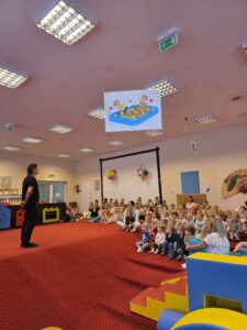 Grupa małych dzieci siedzi blisko siebie na czerwonym dywanie w sali żłobka i z dużym skupieniem ogląda przedstawienie poza kadrem. W tle widać opiekunki siedzące razem z dziećmi. Na twarzach maluchów widać zaciekawienie i zaangażowanie.