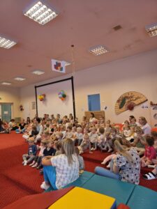 Grupa małych dzieci siedzi blisko siebie na czerwonym dywanie w sali żłobka i z dużym skupieniem ogląda przedstawienie poza kadrem. W tle widać opiekunki siedzące razem z dziećmi. Na twarzach maluchów widać zaciekawienie i zaangażowanie.