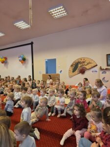 Grupa małych dzieci siedzi blisko siebie na czerwonym dywanie w sali żłobka i z dużym skupieniem ogląda przedstawienie poza kadrem. W tle widać opiekunki siedzące razem z dziećmi. Na twarzach maluchów widać zaciekawienie i zaangażowanie.