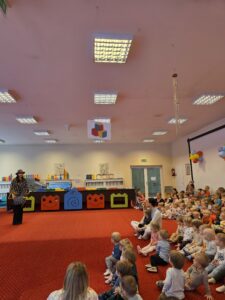 Grupa małych dzieci siedzi blisko siebie na czerwonym dywanie w sali żłobka i z dużym skupieniem ogląda przedstawienie poza kadrem. W tle widać opiekunki siedzące razem z dziećmi. Na twarzach maluchów widać zaciekawienie i zaangażowanie.