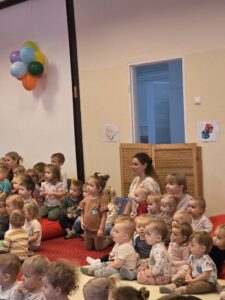 Grupa małych dzieci siedzi blisko siebie na czerwonym dywanie w sali żłobka i z dużym skupieniem ogląda przedstawienie poza kadrem. W tle widać opiekunki siedzące razem z dziećmi. Na twarzach maluchów widać zaciekawienie i zaangażowanie.