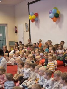 Grupa małych dzieci siedzi blisko siebie na czerwonym dywanie w sali żłobka i z dużym skupieniem ogląda przedstawienie poza kadrem. W tle widać opiekunki siedzące razem z dziećmi. Na twarzach maluchów widać zaciekawienie i zaangażowanie.