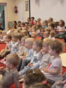 Grupa małych dzieci siedzi blisko siebie na czerwonym dywanie w sali żłobka i z dużym skupieniem ogląda przedstawienie poza kadrem. W tle widać opiekunki siedzące razem z dziećmi. Na twarzach maluchów widać zaciekawienie i zaangażowanie.