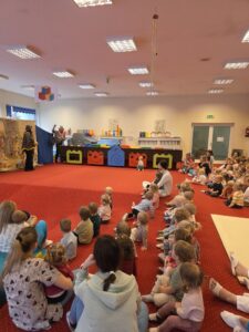 Grupa małych dzieci siedzi blisko siebie na czerwonym dywanie w sali żłobka i z dużym skupieniem ogląda przedstawienie poza kadrem. W tle widać opiekunki siedzące razem z dziećmi. Na twarzach maluchów widać zaciekawienie i zaangażowanie.