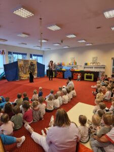 Grupa małych dzieci siedzi blisko siebie na czerwonym dywanie w sali żłobka i z dużym skupieniem ogląda przedstawienie poza kadrem. W tle widać opiekunki siedzące razem z dziećmi. Na twarzach maluchów widać zaciekawienie i zaangażowanie.