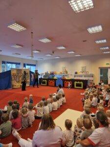 Grupa małych dzieci siedzi blisko siebie na czerwonym dywanie w sali żłobka i z dużym skupieniem ogląda przedstawienie poza kadrem. W tle widać opiekunki siedzące razem z dziećmi. Na twarzach maluchów widać zaciekawienie i zaangażowanie.