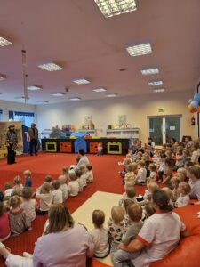 Grupa małych dzieci siedzi blisko siebie na czerwonym dywanie w sali żłobka i z dużym skupieniem ogląda przedstawienie poza kadrem. W tle widać opiekunki siedzące razem z dziećmi. Na twarzach maluchów widać zaciekawienie i zaangażowanie.