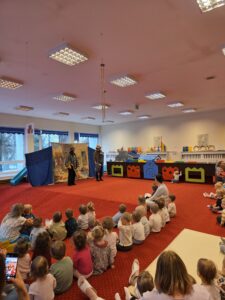 Grupa małych dzieci siedzi blisko siebie na czerwonym dywanie w sali żłobka i z dużym skupieniem ogląda przedstawienie poza kadrem. W tle widać opiekunki siedzące razem z dziećmi. Na twarzach maluchów widać zaciekawienie i zaangażowanie.