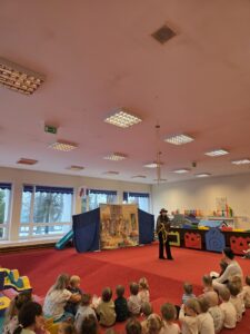 Grupa małych dzieci siedzi blisko siebie na czerwonym dywanie w sali żłobka i z dużym skupieniem ogląda przedstawienie poza kadrem. W tle widać opiekunki siedzące razem z dziećmi. Na twarzach maluchów widać zaciekawienie i zaangażowanie.