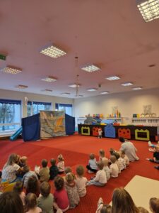 Grupa małych dzieci siedzi blisko siebie na czerwonym dywanie w sali żłobka i z dużym skupieniem ogląda przedstawienie poza kadrem. W tle widać opiekunki siedzące razem z dziećmi. Na twarzach maluchów widać zaciekawienie i zaangażowanie.