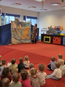 Dzieci siedzące na czerwonym dywanie w sali żłobka oglądają przedstawienie teatralne. Przed nimi ustawiona jest niewielka scena z dekoracją przedstawiającą statek na morzu, zasłonięta po bokach granatowymi kotarami. W tle widać kolorowe meble i zabawki, a opiekunki siedzą razem z dziećmi, wspólnie uczestnicząc w wydarzeniu. Atmosfera skupienia i zaciekawienia.