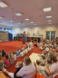 Dzieci siedzące na czerwonym dywanie w sali żłobka oglądają przedstawienie teatralne. Przed nimi ustawiona jest niewielka scena z dekoracją przedstawiającą statek na morzu, zasłonięta po bokach granatowymi kotarami. W tle widać kolorowe meble i zabawki, a opiekunki siedzą razem z dziećmi, wspólnie uczestnicząc w wydarzeniu. Atmosfera skupienia i zaciekawienia.