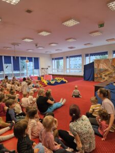 Dzieci siedzące na czerwonym dywanie w sali żłobka oglądają przedstawienie teatralne. Przed nimi ustawiona jest niewielka scena z dekoracją przedstawiającą statek na morzu, zasłonięta po bokach granatowymi kotarami. W tle widać kolorowe meble i zabawki, a opiekunki siedzą razem z dziećmi, wspólnie uczestnicząc w wydarzeniu. Atmosfera skupienia i zaciekawienia.