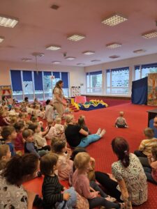 Dzieci siedzące na czerwonym dywanie w sali żłobka oglądają przedstawienie teatralne. Przed nimi ustawiona jest niewielka scena z dekoracją przedstawiającą statek na morzu, zasłonięta po bokach granatowymi kotarami. W tle widać kolorowe meble i zabawki, a opiekunki siedzą razem z dziećmi, wspólnie uczestnicząc w wydarzeniu. Atmosfera skupienia i zaciekawienia.