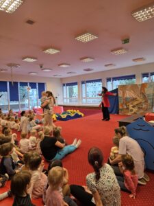 Dzieci siedzące na czerwonym dywanie w sali żłobka oglądają przedstawienie teatralne. Przed nimi ustawiona jest niewielka scena z dekoracją przedstawiającą statek na morzu, zasłonięta po bokach granatowymi kotarami. W tle widać kolorowe meble i zabawki, a opiekunki siedzą razem z dziećmi, wspólnie uczestnicząc w wydarzeniu. Atmosfera skupienia i zaciekawienia.