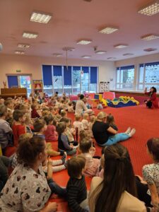 Dzieci siedzące na czerwonym dywanie w sali żłobka oglądają przedstawienie teatralne. Przed nimi ustawiona jest niewielka scena z dekoracją przedstawiającą statek na morzu, zasłonięta po bokach granatowymi kotarami. W tle widać kolorowe meble i zabawki, a opiekunki siedzą razem z dziećmi, wspólnie uczestnicząc w wydarzeniu. Atmosfera skupienia i zaciekawienia.