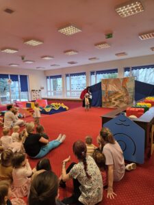 Dzieci siedzące na czerwonym dywanie w sali żłobka oglądają przedstawienie teatralne. Przed nimi ustawiona jest niewielka scena z dekoracją przedstawiającą statek na morzu, zasłonięta po bokach granatowymi kotarami. W tle widać kolorowe meble i zabawki, a opiekunki siedzą razem z dziećmi, wspólnie uczestnicząc w wydarzeniu. Atmosfera skupienia i zaciekawienia.