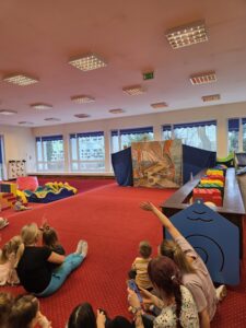 Dzieci siedzące na czerwonym dywanie w sali żłobka oglądają przedstawienie teatralne. Przed nimi ustawiona jest niewielka scena z dekoracją przedstawiającą statek na morzu, zasłonięta po bokach granatowymi kotarami. W tle widać kolorowe meble i zabawki, a opiekunki siedzą razem z dziećmi, wspólnie uczestnicząc w wydarzeniu. Atmosfera skupienia i zaciekawienia.