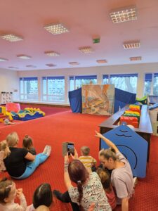 Dzieci siedzące na czerwonym dywanie w sali żłobka oglądają przedstawienie teatralne. Przed nimi ustawiona jest niewielka scena z dekoracją przedstawiającą statek na morzu, zasłonięta po bokach granatowymi kotarami. W tle widać kolorowe meble i zabawki, a opiekunki siedzą razem z dziećmi, wspólnie uczestnicząc w wydarzeniu. Atmosfera skupienia i zaciekawienia.