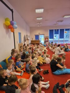 Dzieci siedzące na czerwonym dywanie w sali żłobka oglądają przedstawienie teatralne. Przed nimi ustawiona jest niewielka scena z dekoracją przedstawiającą statek na morzu, zasłonięta po bokach granatowymi kotarami. W tle widać kolorowe meble i zabawki, a opiekunki siedzą razem z dziećmi, wspólnie uczestnicząc w wydarzeniu. Atmosfera skupienia i zaciekawienia.