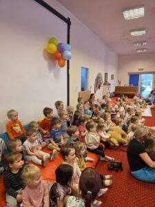 Dzieci siedzące na czerwonym dywanie w sali żłobka oglądają przedstawienie teatralne. Przed nimi ustawiona jest niewielka scena z dekoracją przedstawiającą statek na morzu, zasłonięta po bokach granatowymi kotarami. W tle widać kolorowe meble i zabawki, a opiekunki siedzą razem z dziećmi, wspólnie uczestnicząc w wydarzeniu. Atmosfera skupienia i zaciekawienia.