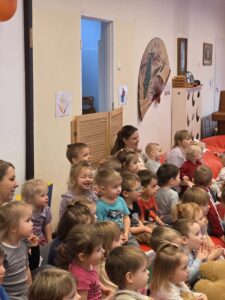 Dzieci siedzące na czerwonym dywanie w sali żłobka oglądają przedstawienie teatralne. Przed nimi ustawiona jest niewielka scena z dekoracją przedstawiającą statek na morzu, zasłonięta po bokach granatowymi kotarami. W tle widać kolorowe meble i zabawki, a opiekunki siedzą razem z dziećmi, wspólnie uczestnicząc w wydarzeniu. Atmosfera skupienia i zaciekawienia.