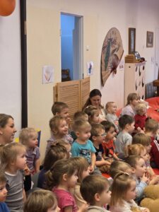 Dzieci siedzące na czerwonym dywanie w sali żłobka oglądają przedstawienie teatralne. Przed nimi ustawiona jest niewielka scena z dekoracją przedstawiającą statek na morzu, zasłonięta po bokach granatowymi kotarami. W tle widać kolorowe meble i zabawki, a opiekunki siedzą razem z dziećmi, wspólnie uczestnicząc w wydarzeniu. Atmosfera skupienia i zaciekawienia.