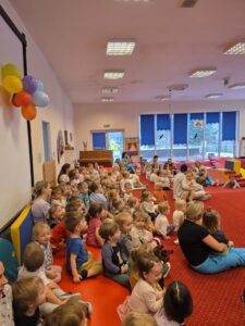 Dzieci siedzące na czerwonym dywanie w sali żłobka oglądają przedstawienie teatralne. Przed nimi ustawiona jest niewielka scena z dekoracją przedstawiającą statek na morzu, zasłonięta po bokach granatowymi kotarami. W tle widać kolorowe meble i zabawki, a opiekunki siedzą razem z dziećmi, wspólnie uczestnicząc w wydarzeniu. Atmosfera skupienia i zaciekawienia.