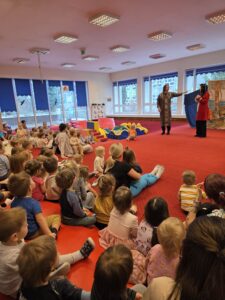 Dzieci siedzące na czerwonym dywanie w sali żłobka oglądają przedstawienie teatralne. Przed nimi ustawiona jest niewielka scena z dekoracją przedstawiającą statek na morzu, zasłonięta po bokach granatowymi kotarami. W tle widać kolorowe meble i zabawki, a opiekunki siedzą razem z dziećmi, wspólnie uczestnicząc w wydarzeniu. Atmosfera skupienia i zaciekawienia.