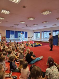 Dzieci siedzące na czerwonym dywanie w sali żłobka oglądają przedstawienie teatralne. Przed nimi ustawiona jest niewielka scena z dekoracją przedstawiającą statek na morzu, zasłonięta po bokach granatowymi kotarami. W tle widać kolorowe meble i zabawki, a opiekunki siedzą razem z dziećmi, wspólnie uczestnicząc w wydarzeniu. Atmosfera skupienia i zaciekawienia.