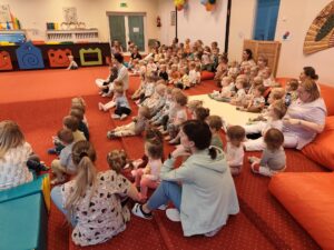 Grupa małych dzieci siedzi blisko siebie na czerwonym dywanie w sali żłobka i z dużym skupieniem ogląda przedstawienie poza kadrem. W tle widać opiekunki siedzące razem z dziećmi. Na twarzach maluchów widać zaciekawienie i zaangażowanie.