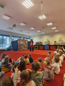 Grupa małych dzieci siedzi blisko siebie na czerwonym dywanie w sali żłobka i z dużym skupieniem ogląda przedstawienie poza kadrem. W tle widać opiekunki siedzące razem z dziećmi. Na twarzach maluchów widać zaciekawienie i zaangażowanie.