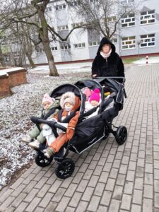 Opiekunka żłobka prowadząca wieloosobowy wózek spacerowy z dziećmi podczas zimowego spaceru na terenie placówki