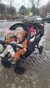 Opiekunka żłobka prowadząca wieloosobowy wózek spacerowy z dziećmi podczas zimowego spaceru na terenie placówki