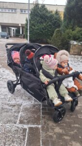Opiekunka żłobka prowadząca wieloosobowy wózek spacerowy z dziećmi podczas zimowego spaceru na terenie placówki